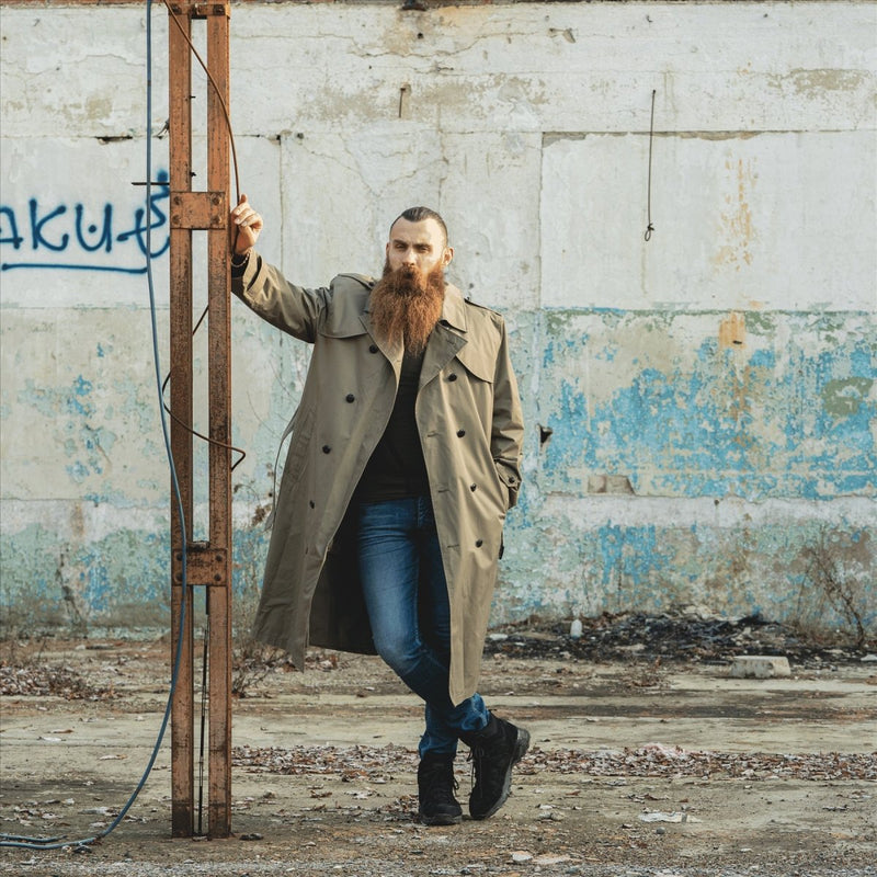 Man wearing a genuine Dutch army khaki long officer trench coat standing outdoors by a rusty metal structure