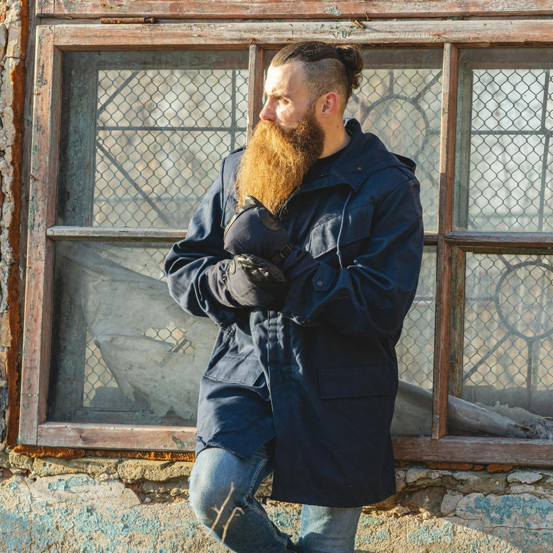 Man wearing a blue Dutch military officer parka with hood, standing in front of a weathered window outdoors.