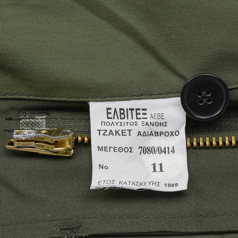 Close-up of a white label with Greek text sewn inside an olive green military field jacket near the zipper and button.