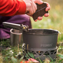 Person breaking chocolate over Primus PrimeTech 1.3L camping stove pot outdoors on grass during cooking